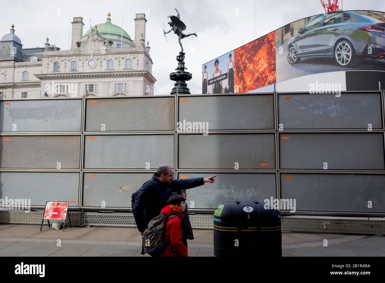 An adult points out a London site next to a temporary construction hoarding beneath the partially hidden statue of the world famous London Victorian-era landmark, Eros in Piccadilly Circus, on 25th February 2020, in London, England. Eros, or the Shaftesbury Memorial Fountain is located at the southeastern side of Piccadilly Circus in London, United Kingdom. Moved after World War II from its original position in the centre, it was erected in 1892–1893 to commemorate the philanthropic works of Lord Shaftesbury, who was a famous Victorian politician and philanthropist. The monument is surmounted Stock Photo