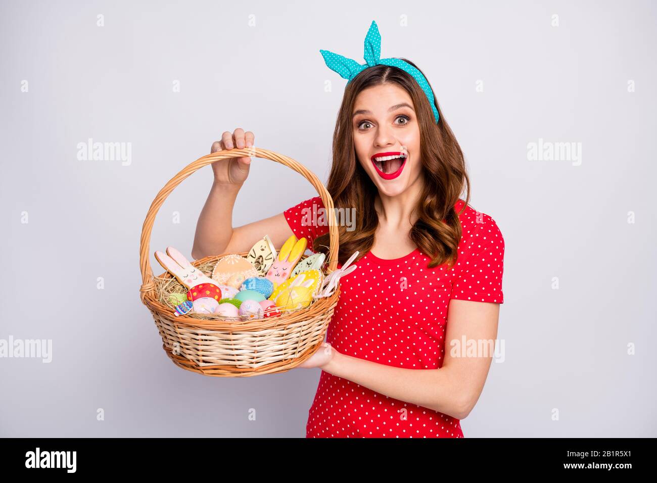 Shocked surprised redhair girl hold big wicker basket painted colorful blue eggs sweet easter confectionery shaped bunny wear red t-shirt retro style Stock Photo