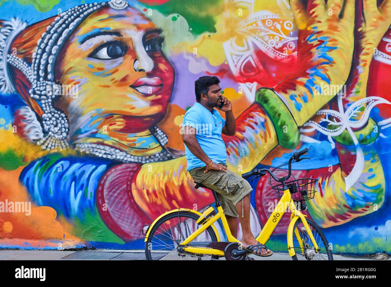 An Indian 'foreign worker' making a call on his bicycle, in front a colorful wall painting depicting a South Indian dancer, in Little India, Singapore Stock Photo