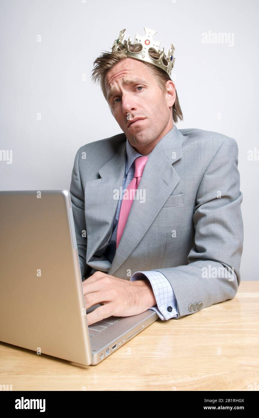 Smug businessman wearing royal gold crown sitting in front of his laptop computer Stock Photo
