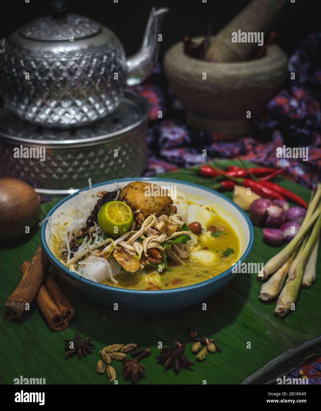 Bowl of traditional Malaysian soto or broth with chicken, noodles and vegetables garnished with lime surrounded by ingredients Stock Photo