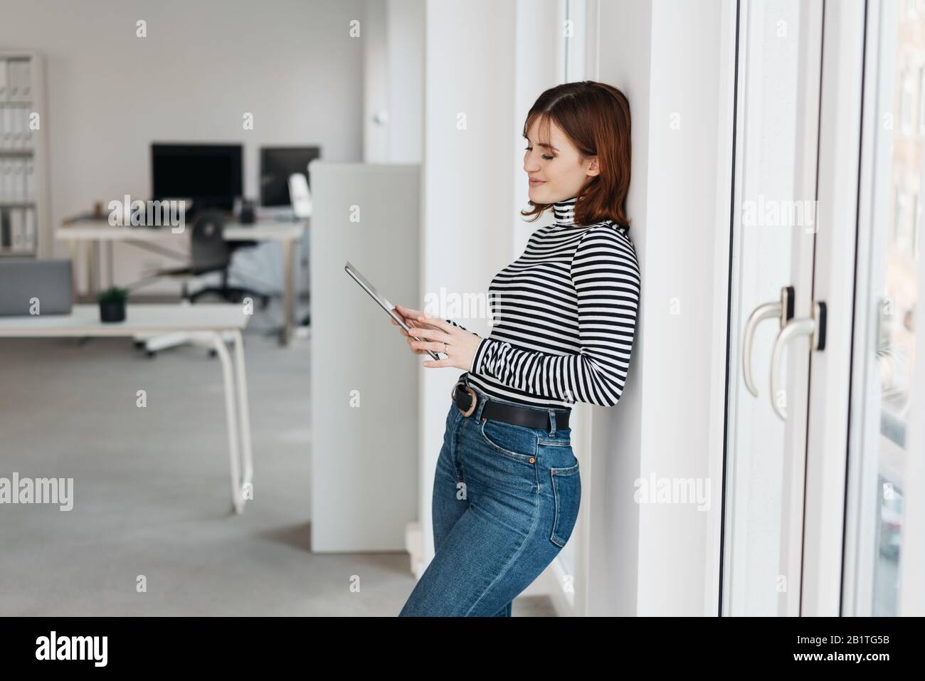 Beautiful young woman in stripped shirt and jeans standing with tablet pc  in the office, leaning back on white wall and looking at the screen of the d  Stock Photo - Alamy
