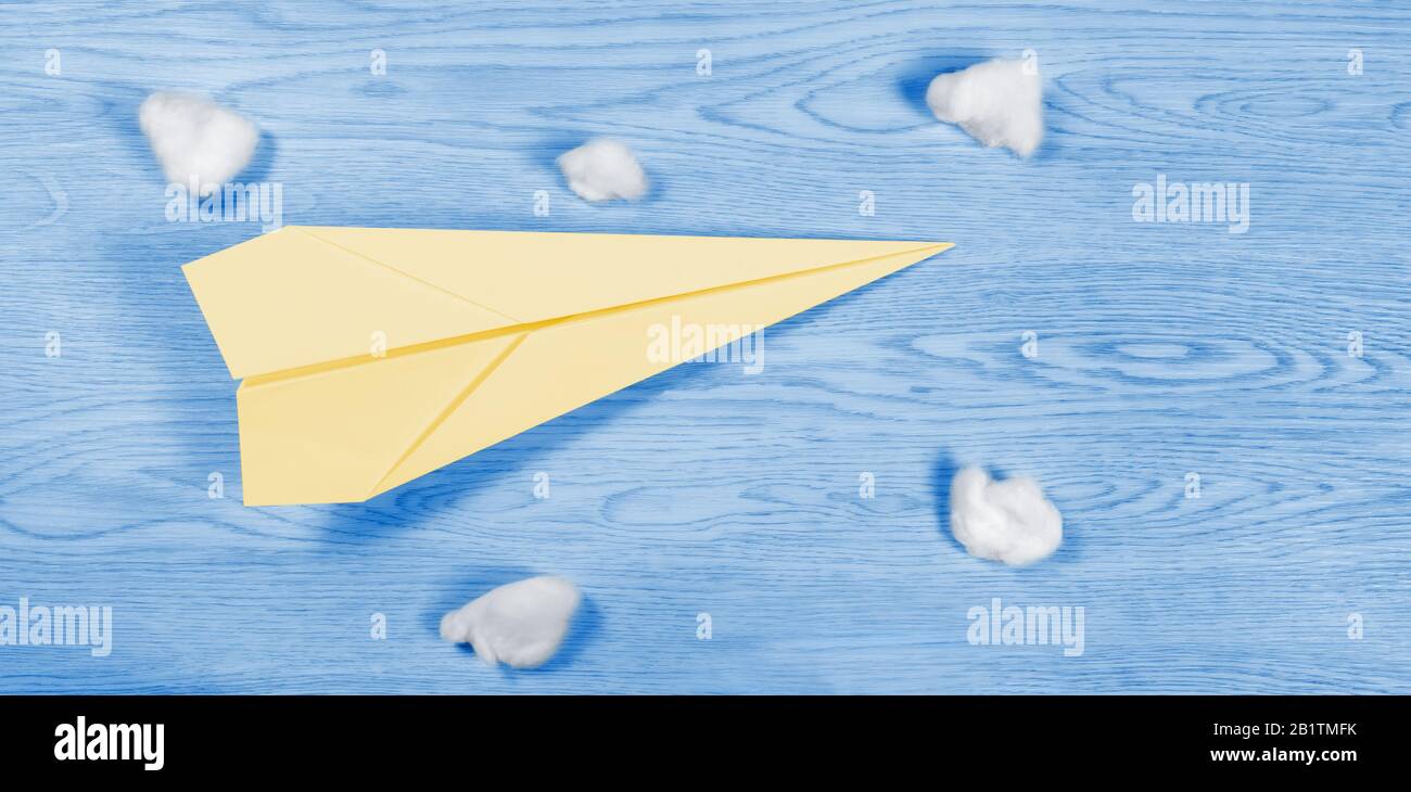 Yellow paper airplane among cotton wool balls over a wooden blue background resembling a blue sky and a few clouds Stock Photo