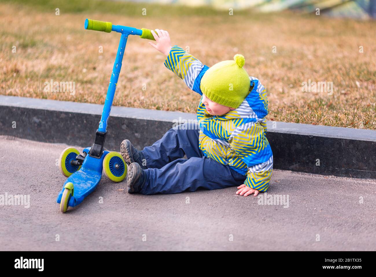 Cute little boy falling off his scooter . Kid getting hurt while riding a kick scooter. Falling scooter accident. Boy trying to stand up Stock Photo