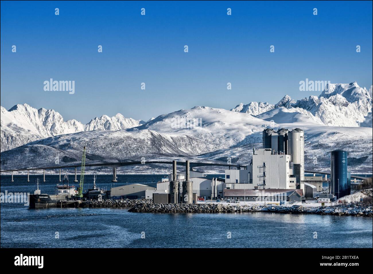 Factory in Norway with a moored barge, a bridge and snow covered mountains in the background. Stock Photo