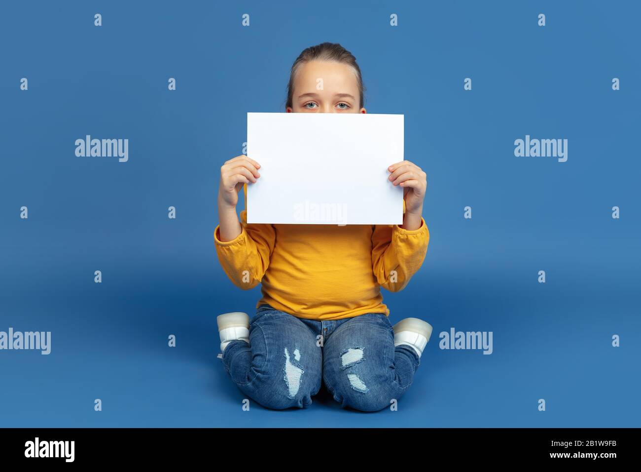 Portrait of sad little girl sitting isolated on blue studio background. How it feels to be autist. Modern problems, new vision of social issues. Concept of autism, childhood, healthcare, medicine. Stock Photo