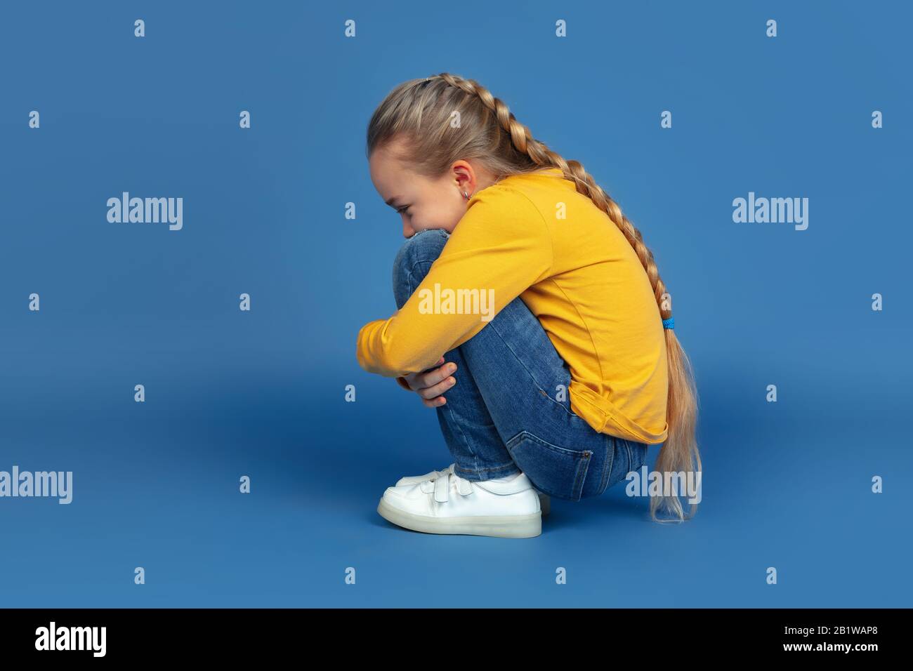 Portrait of sad little girl sitting isolated on blue studio background. How it feels to be autist. Modern problems, new vision of social issues. Concept of autism, childhood, healthcare, medicine. Stock Photo