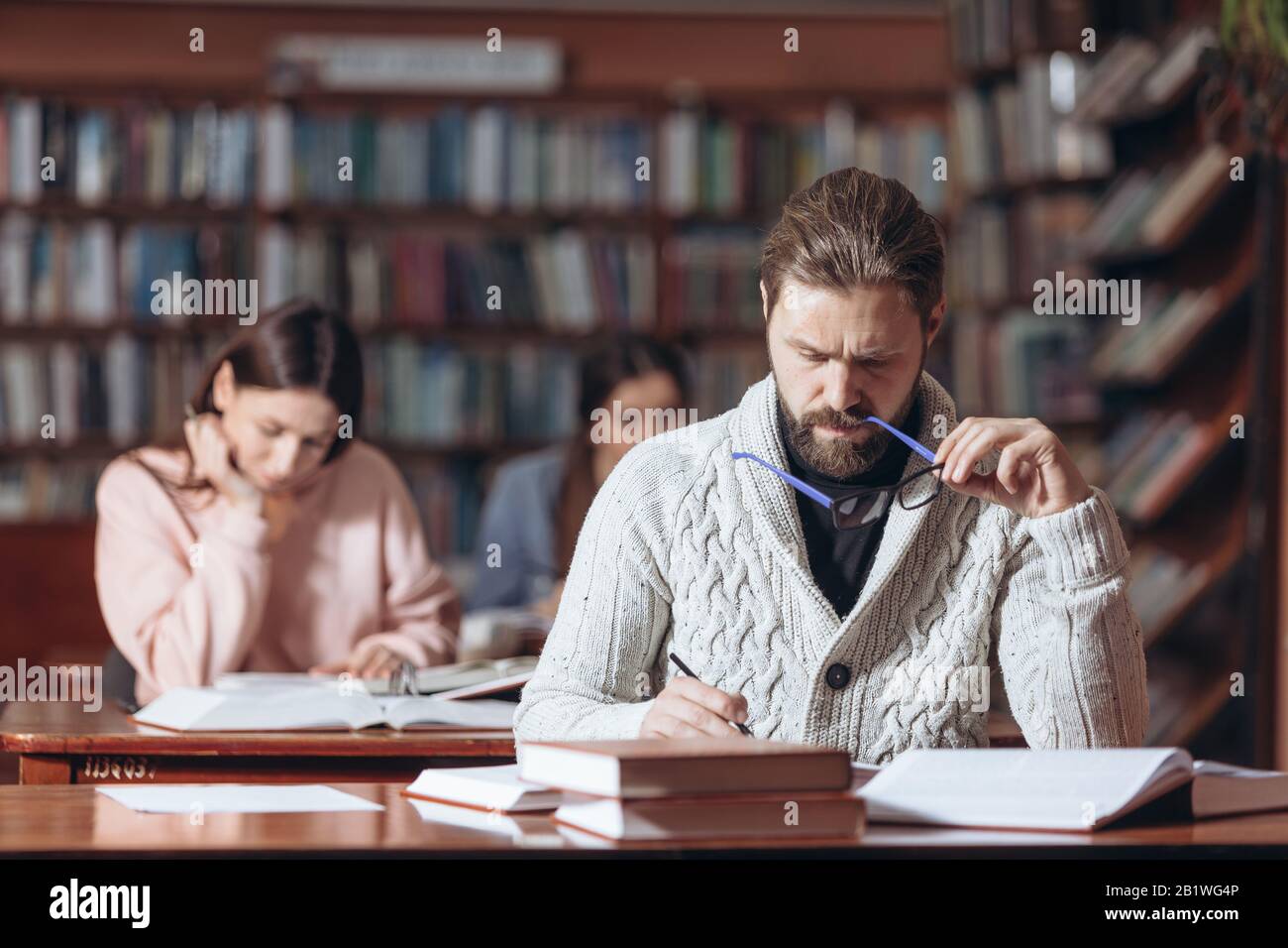 Concentrated unshaven man in knitted sweater and eyeglasses taking notes while reading books at public library. Mature male sitting at table with blur Stock Photo