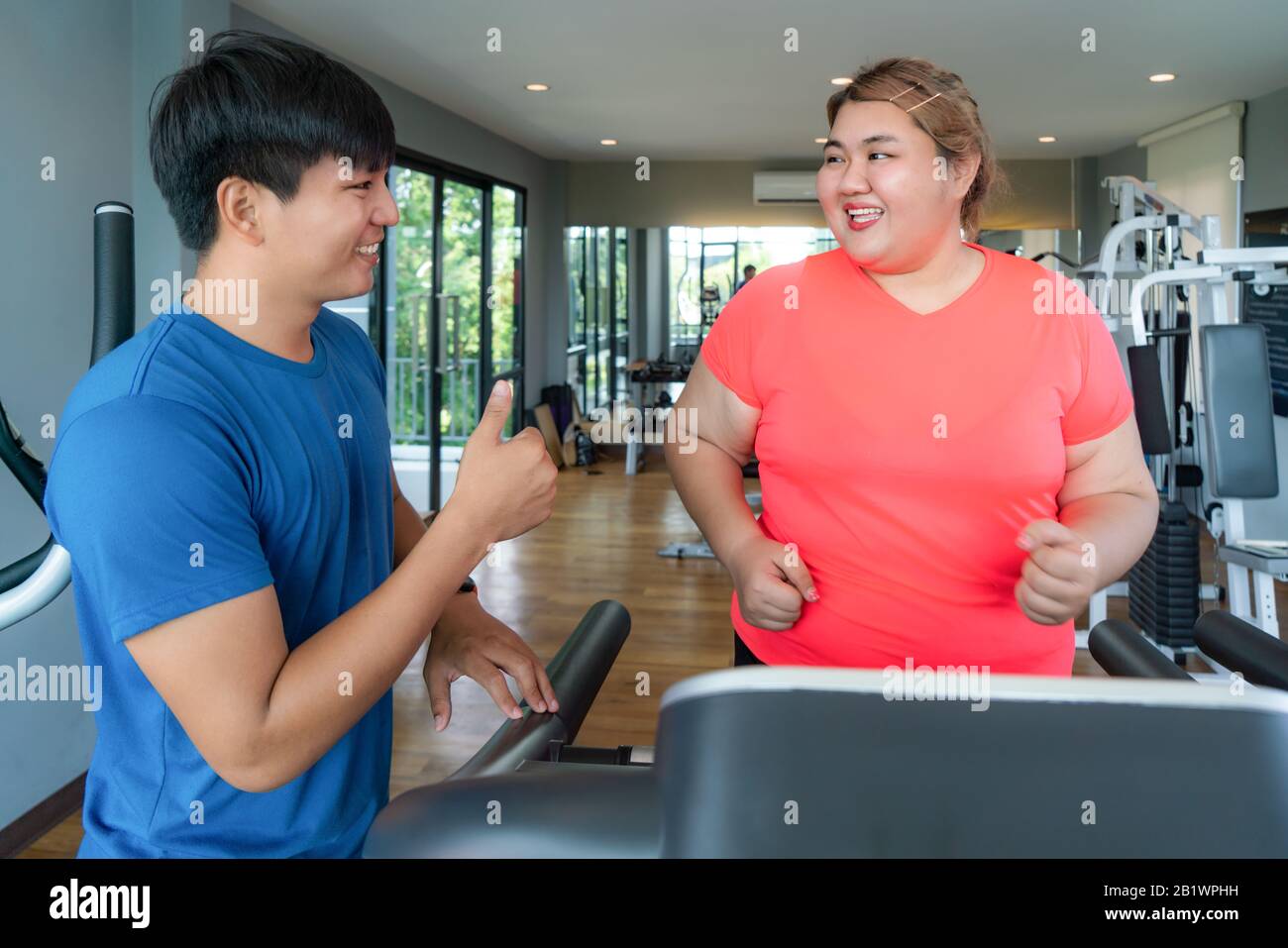 Two Asian trainer man and Overweight woman exercising training on treadmill in gym, trainer looking happy her result and thumb up during workout. Fat Stock Photo
