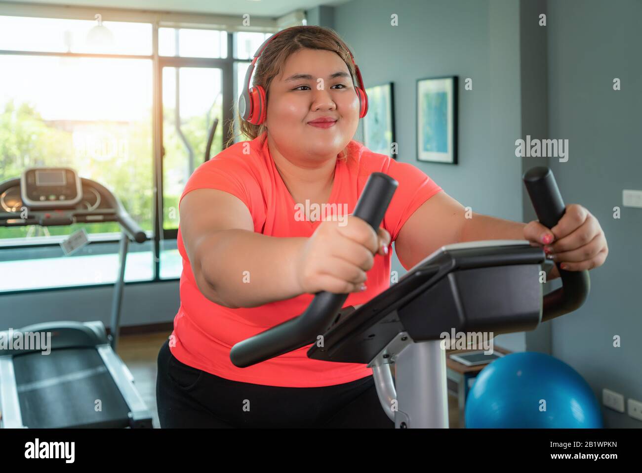 Happy Asian Overweight woman with earphone training on exercise bike in modern gym, happy and smile during workout. Fat woman take care of health and Stock Photo