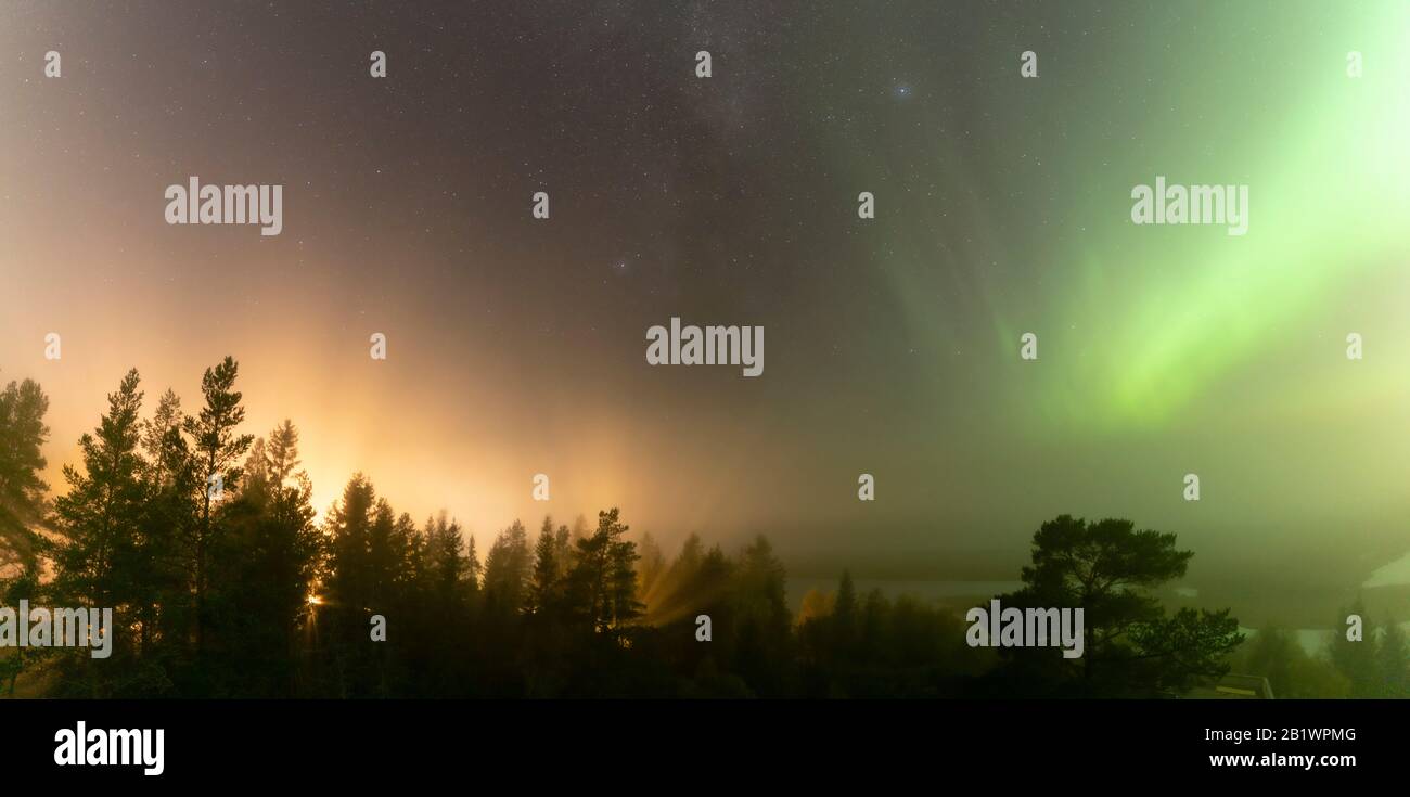 Panoramic view of brilliant green Aurora shining over Swedish foggy forest landscape, light rays from a village and Northern Lights color sky in diffe Stock Photo
