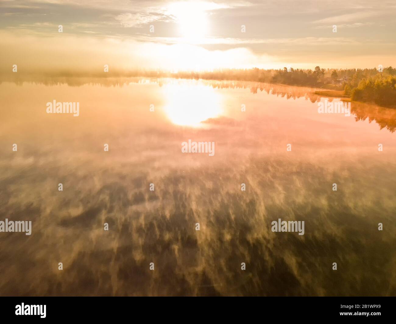 Beautiful aerial photo above summer lake with fog laying above water surface and sunrise over forest landscape and village, still air. Red orange Sun Stock Photo