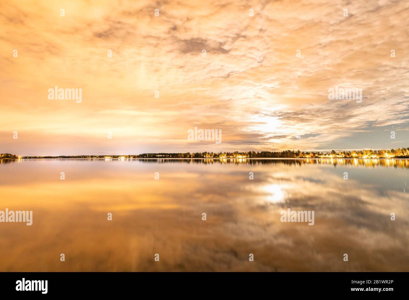 Night photo of clouds mirror reflected at Swedish countryside lake, moon shines behind clouds and gives unreal colors Stock Photo