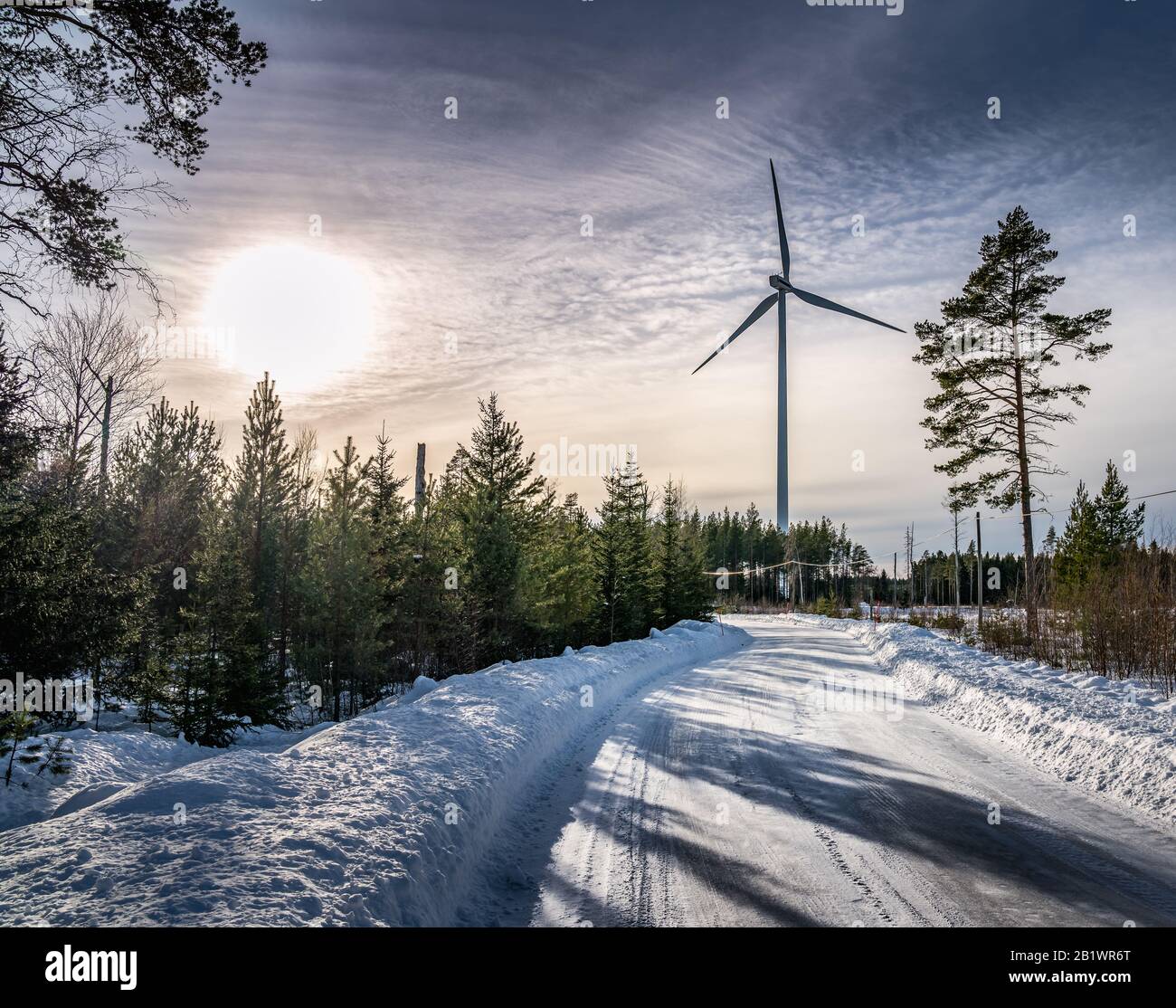 photo of windmill and curvy road in winter pine tree forest, blue sky, foggy winter Sun, Sweden, environment friendly green energy Stock Photo