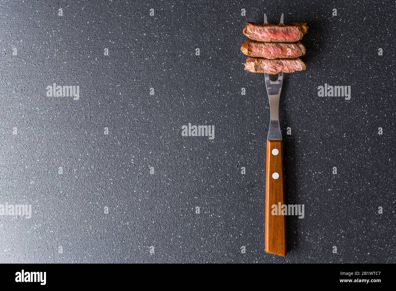 New york strip loin beef steak on a meat fork against black stone background. Stock Photo