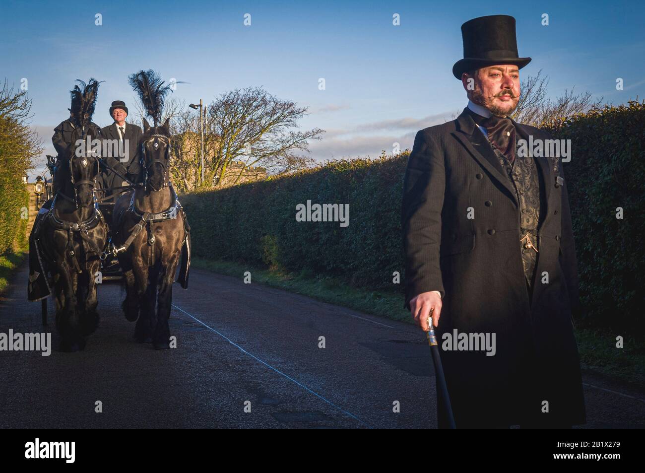 A funeral director, dressed in traditional formal morning dress with top hat, and cane, leads a horse drawn hearse as part of a funeral cortege. Stock Photo