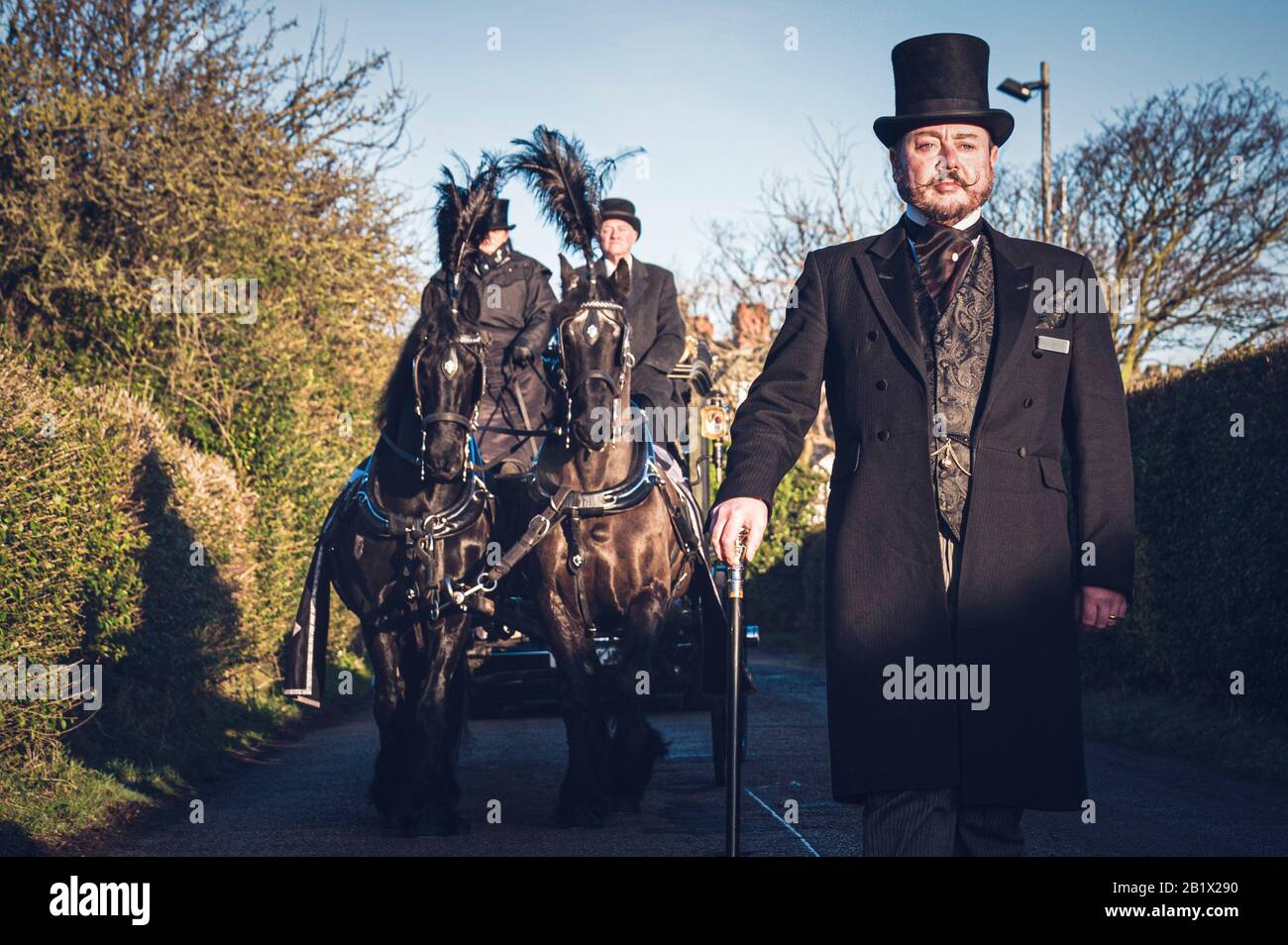 A funeral director, dressed in traditional formal morning dress with top hat, and cane, leads a horse drawn hearse as part of a funeral cortege. Stock Photo