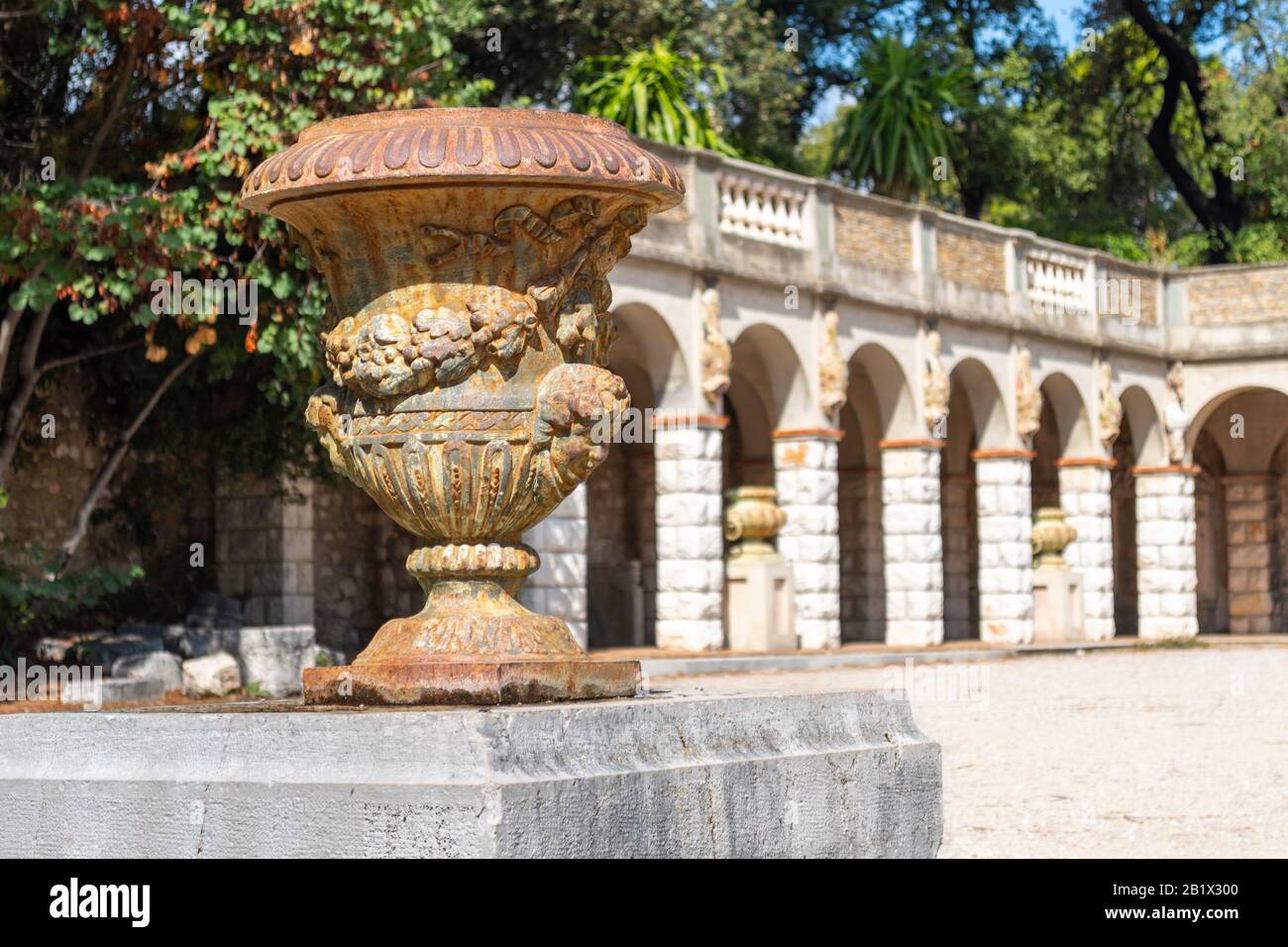 An ornate antique vase sits atop a column with a historic arched building blurred behind on Castle Hill in Nice, France. Stock Photo
