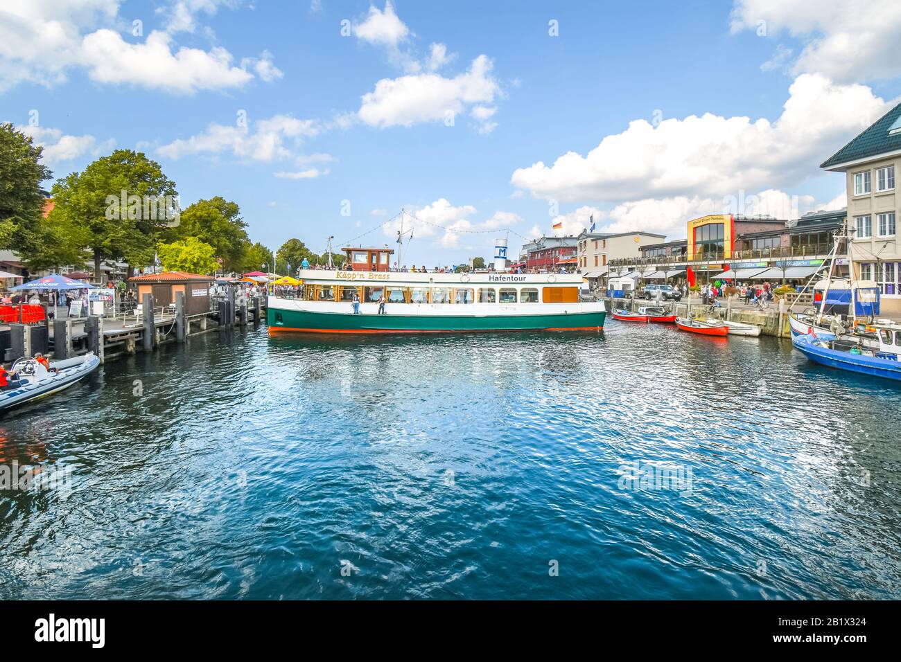 A tourist cruise boat turns sideways in the Alter Strom canal alongside shops and fishing boats on the Baltic Sea resort. Stock Photo
