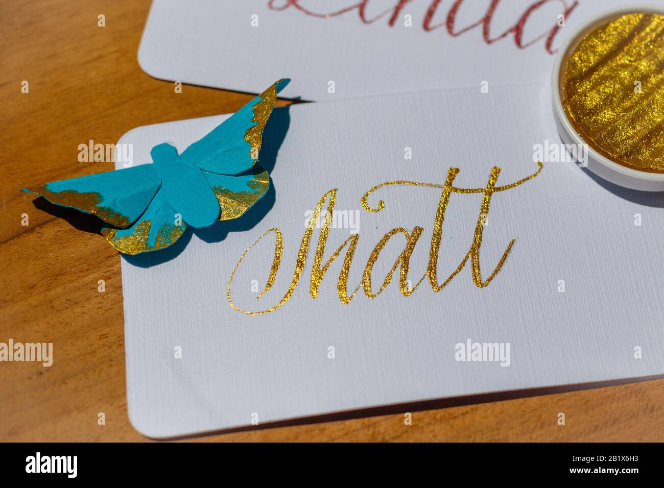 Wedding calligraphy - handwritten place cards with names and paper butterflies. Golden writing on white paper. Stock Photo