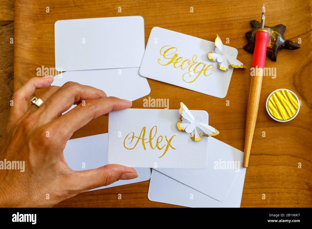 Wedding calligraphy - female hand holding handwritten place cards with names and paper butterflies. Golden writing on white paper. Stock Photo