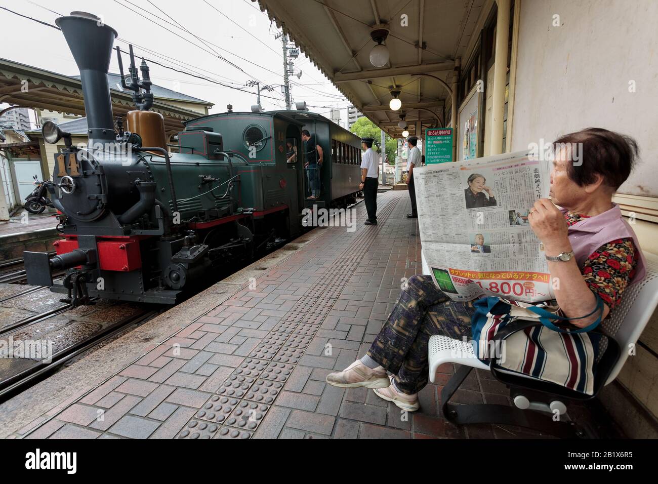 An older Japanese woman reads a newspaper at a station with the famous Botchan Ressha train behind Matsuyama, Eihime, Japan.. Stock Photo