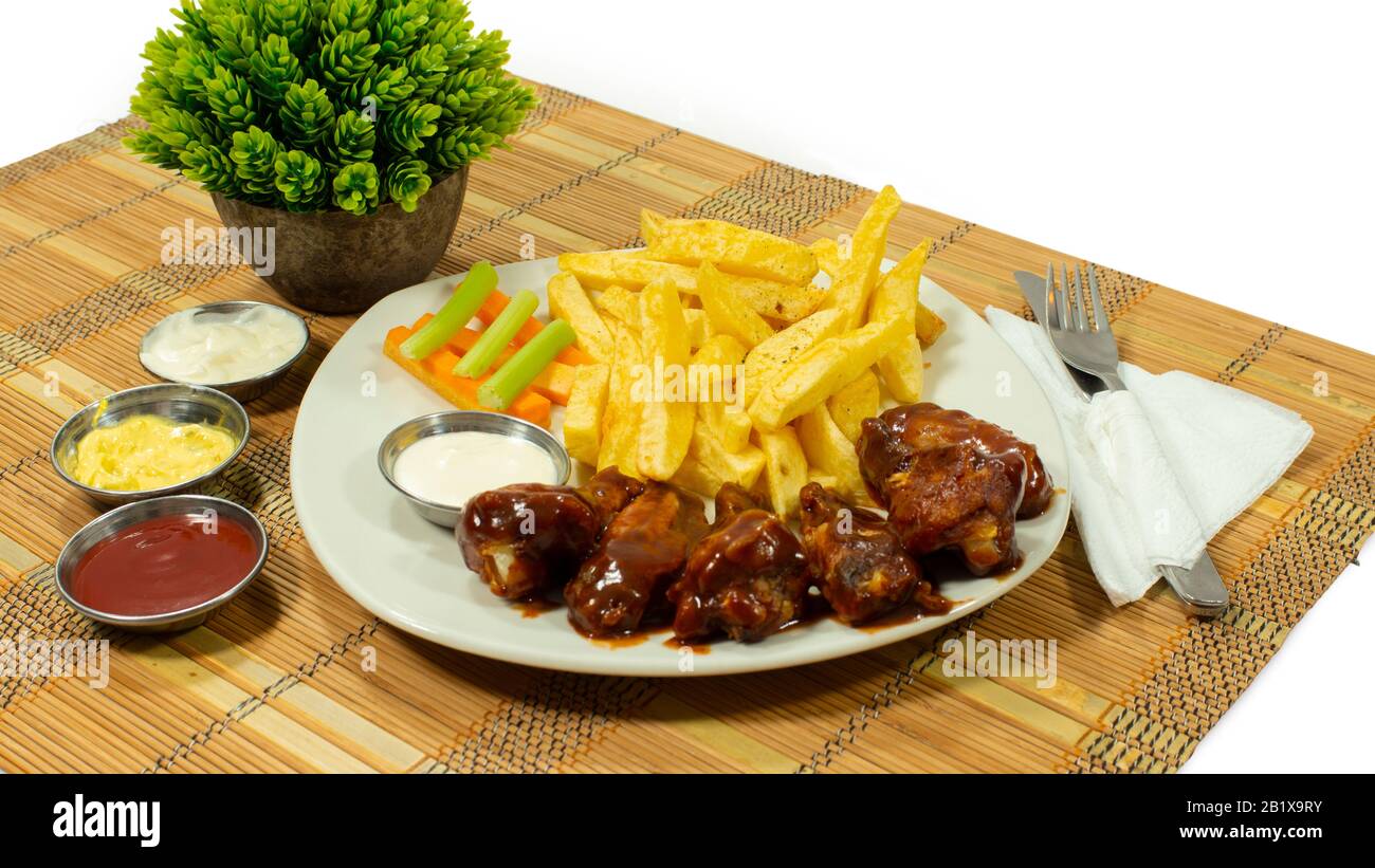 White plate with BBQ chicken wings accompanied by french fries, carrot and celery pieces, with sauces in small plates on white background Stock Photo
