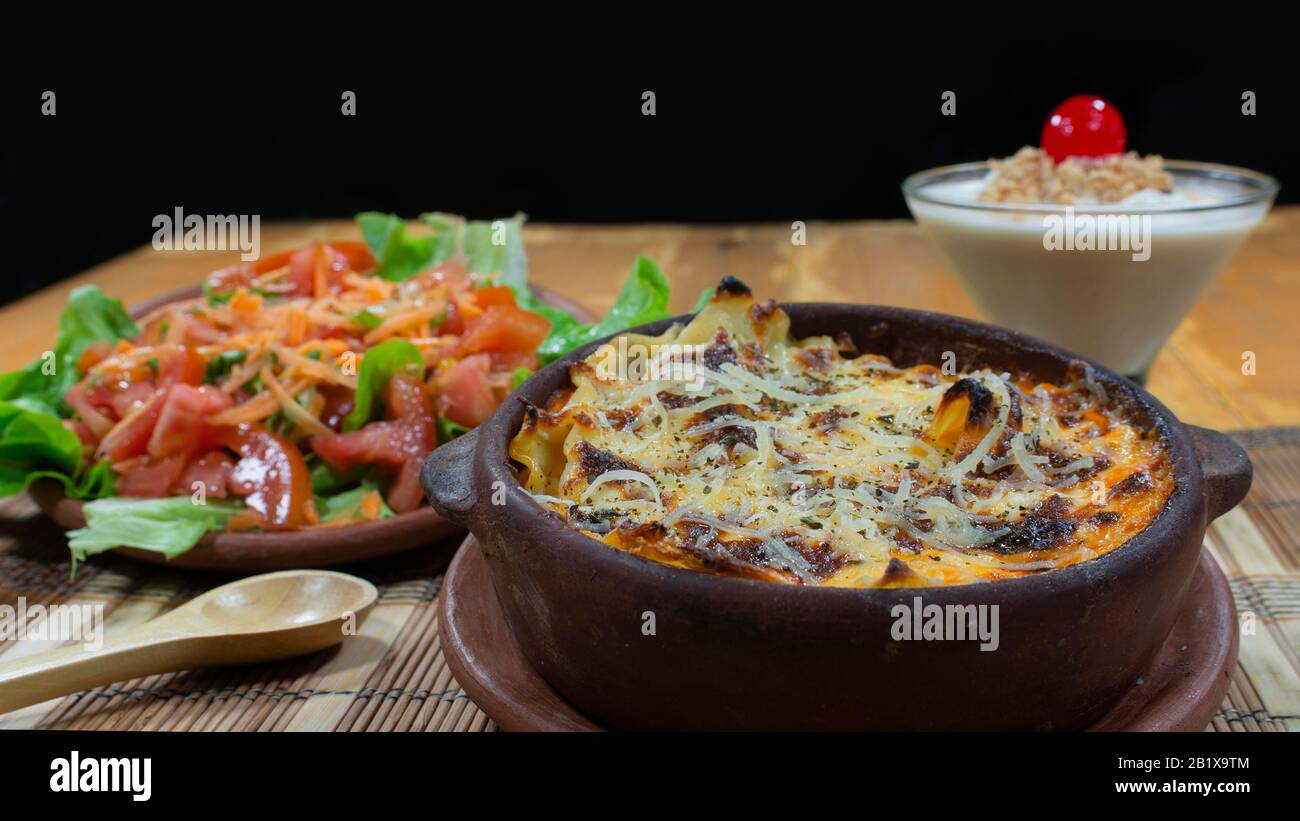 Clay dish with meat lasagna accompanied by carrot, tomato and lettuce salad on a wooden table on black background Stock Photo