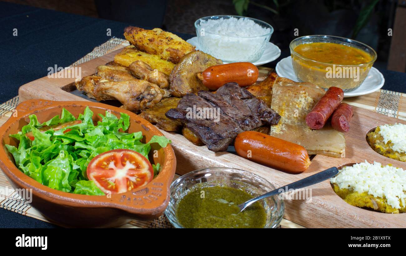 Large wooden tray with grilled meats and sausages accompanied by sauces, fried cassava, a plate of rice and tomato and lettuce salad on a black table Stock Photo