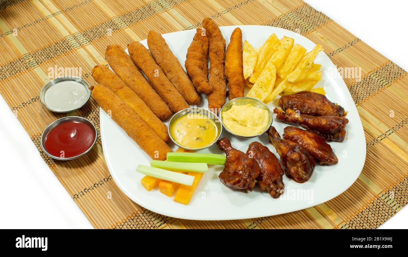 White plate with BBQ chicken wings and breaded chicken fingers accompanied by french fries, carrot and celery pieces, with sauces in small plates on w Stock Photo