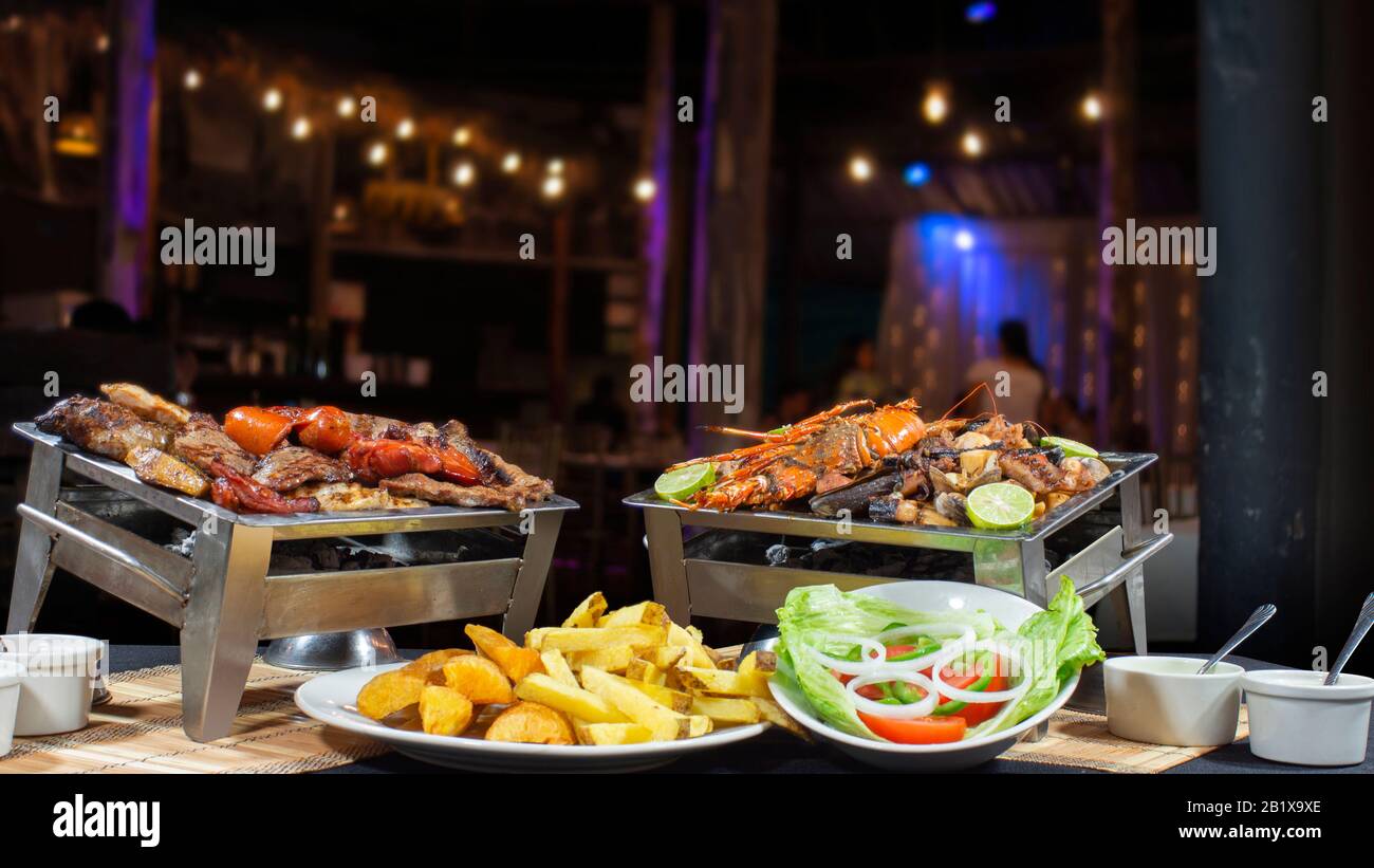 Grilled meat with sausages and seafood grill with lobster, shrimp, shell, clams accompanied by salad and fries with unfocused restaurant background Stock Photo