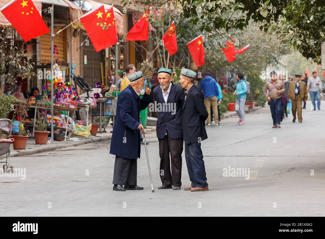 Three elderly men of the Uyghur minority having a conversation at a street in Kashgar Old Town. Chinese flags are mounted on the house in the back. Stock Photo