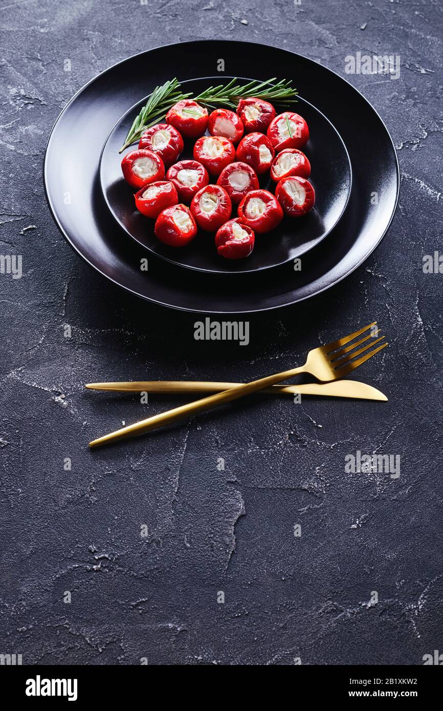 Party food ricotta stuffed hot cherry peppers served on a black plate with rosemary sprigs and golden cutlery on a dark concrete background, vertical Stock Photo