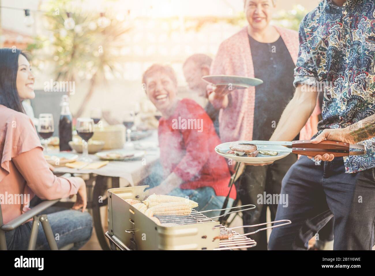 Trendy man cooking and serving meat at barbecue dinner outdoor - Chef grilling food for family friends at bbq meal outside - Summer lifestyle, friends Stock Photo