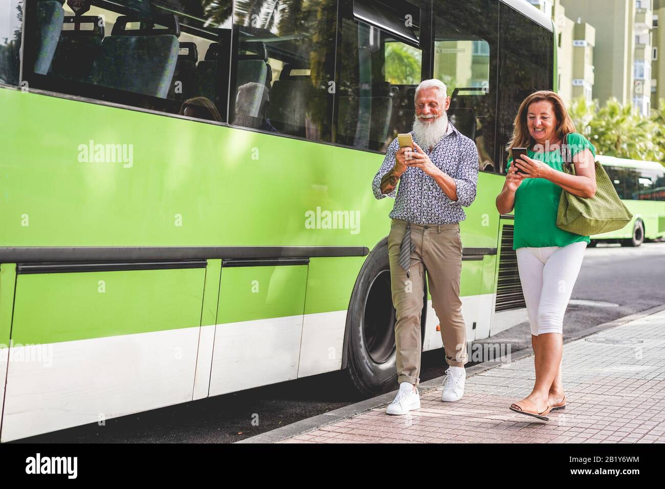 Happy senior couple using smartphones at bus station - 60's age people having fun with technology trends - Transport and joyful elderly concept - Focu Stock Photo