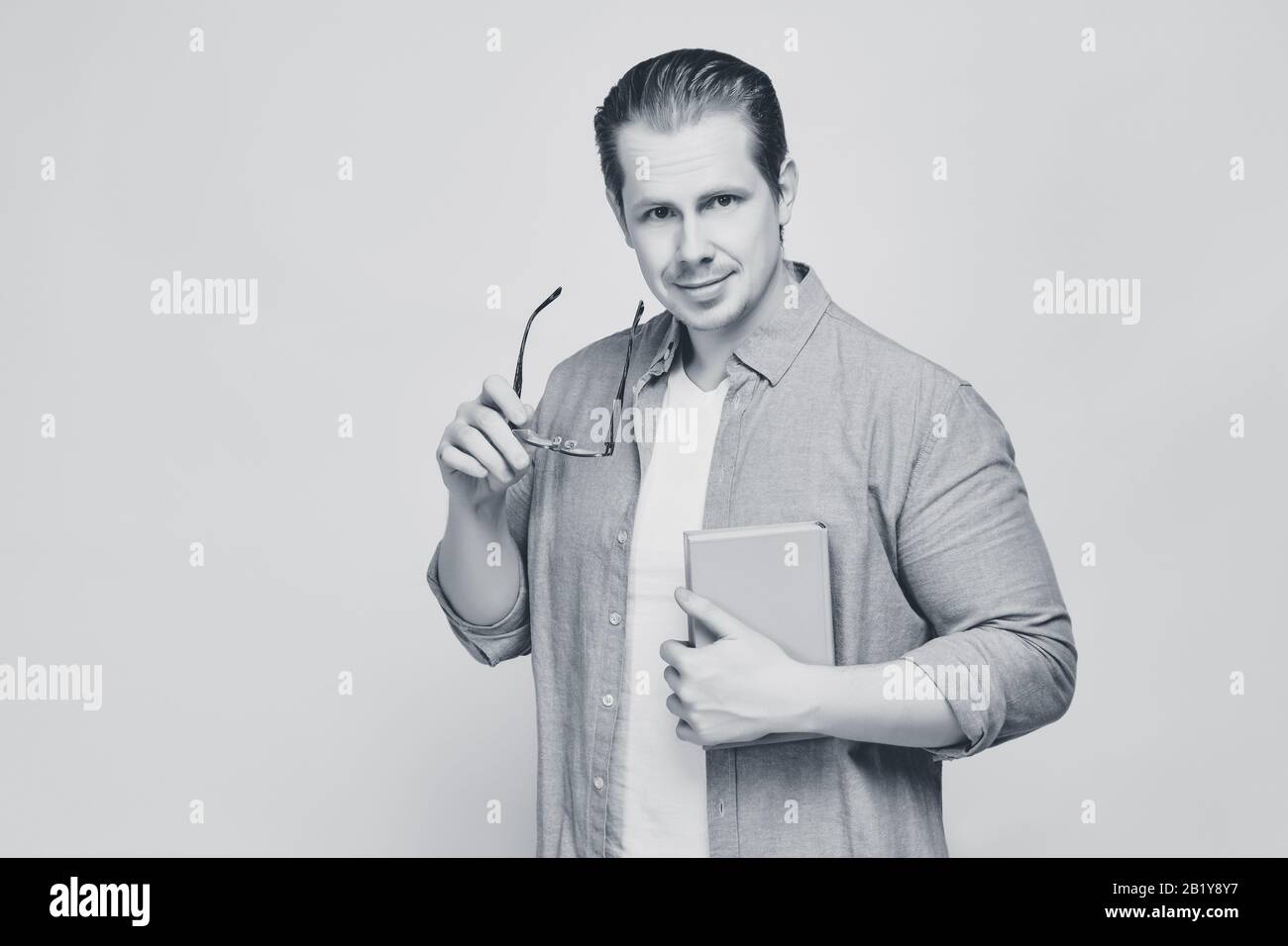 A man in a shirt on a yellow background holds a book while taking off his glasses and looks at the camera. Black and white. Close up. Stock Photo