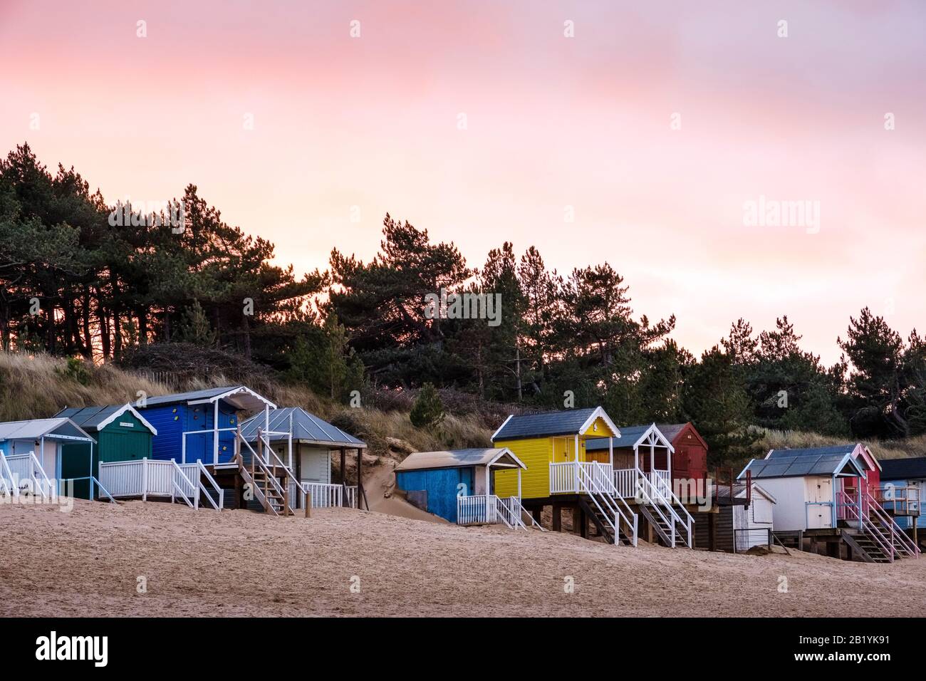 The iconic, brightly coloured beach huts on Wells beach, at sunset in winter, with the pine woods behind. Stock Photo