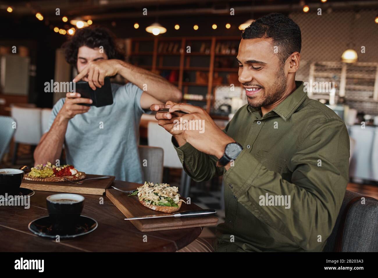 Young afro-americna man with caucasian friend clicking photo of italian bruschetta with coffee in cafe while enjoying lunch Stock Photo