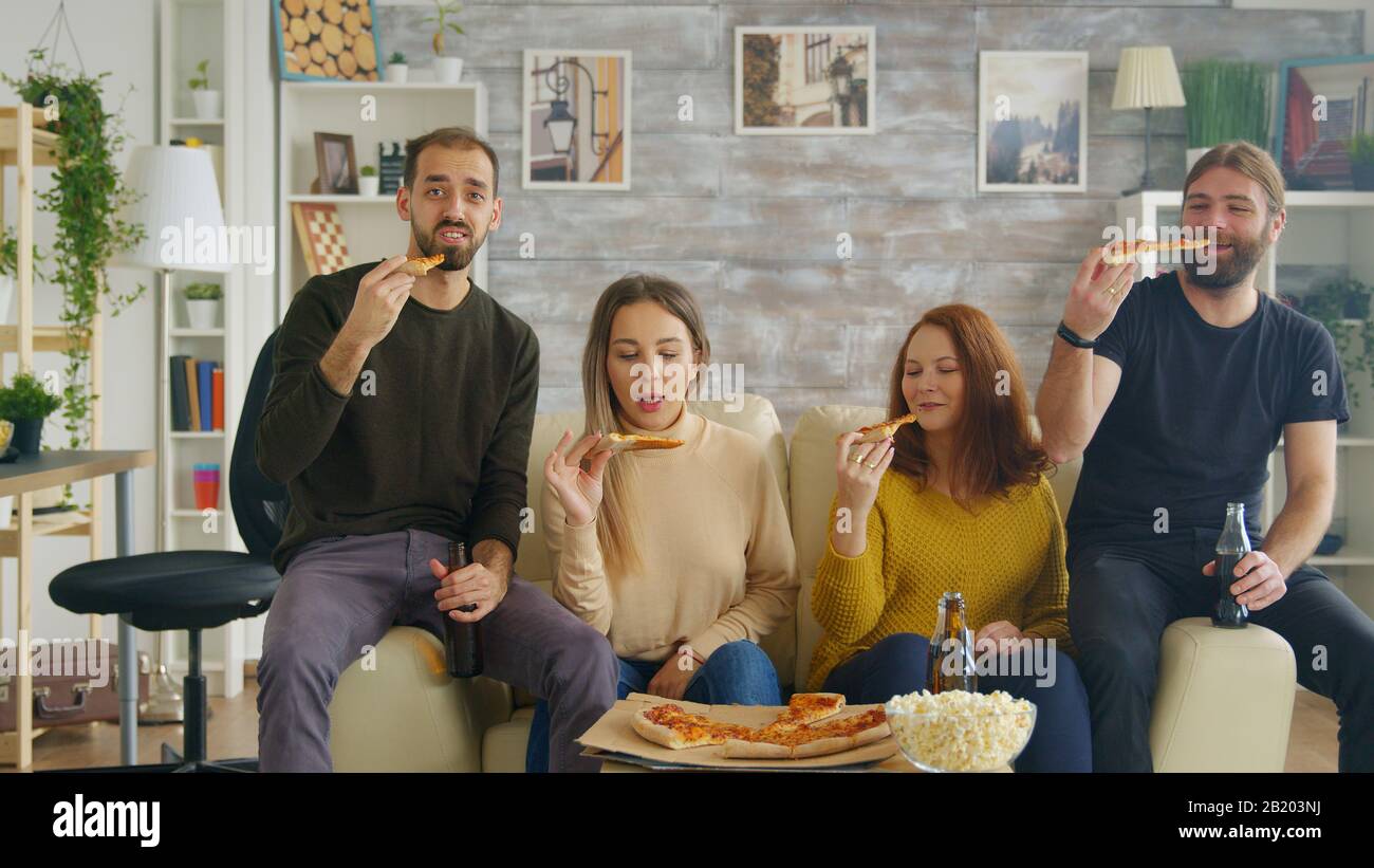 Man changing channels on tv using remote control while a tv show with his friends, drinking beer and eating pizza. Stock Photo