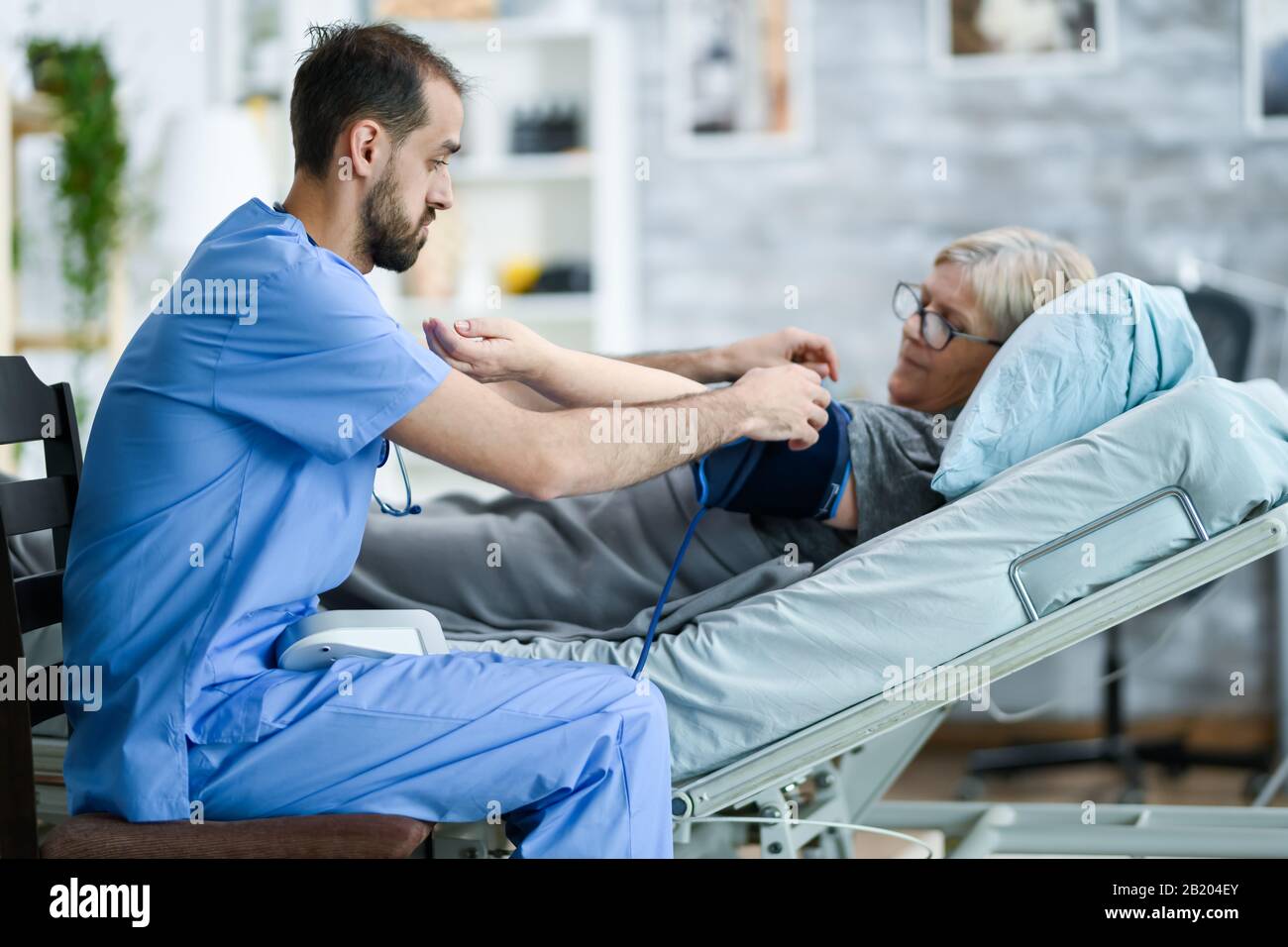 Male caretaker in a nursing home taking bood pressure of old woman laying in bed. Stock Photo