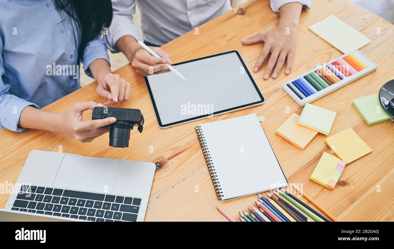 Cropped shot of young designer team sitting at the wooden working desk while holding camera in hands and discussing about information in tablet. Brain Stock Photo
