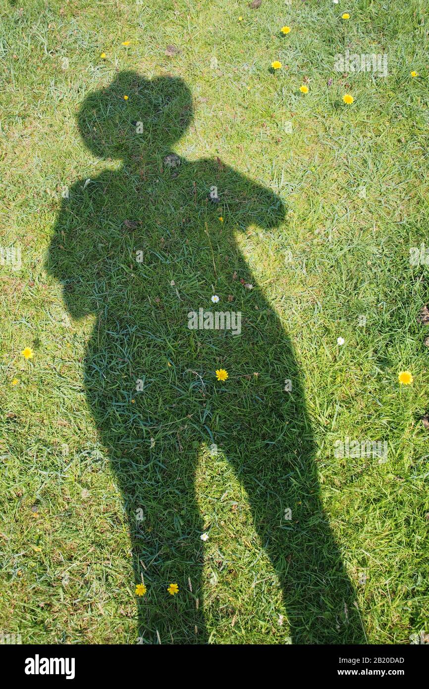 A shadow of a standing woman on grass with dandelions, wearing trousers and a wide brimmed hat Stock Photo
