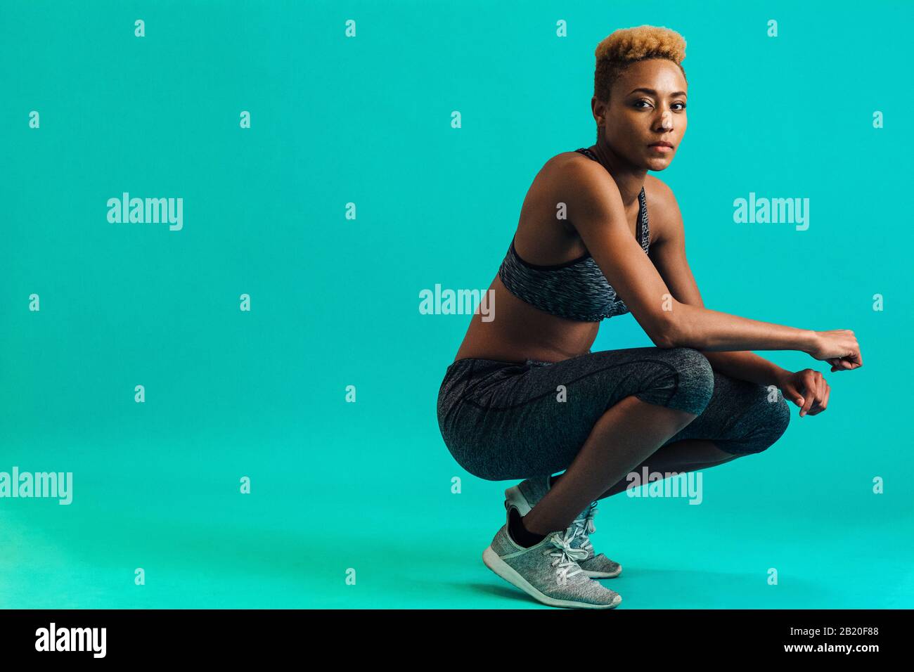 Portrait of a serious female athlete in sports bra and gym tights, squatting and looking at camera, against studio background Stock Photo