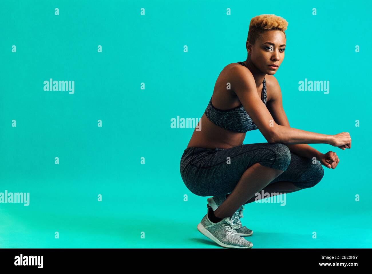 Portrait of a serious female athlete in sports bra and gym tights, squatting and looking off, against studio background Stock Photo