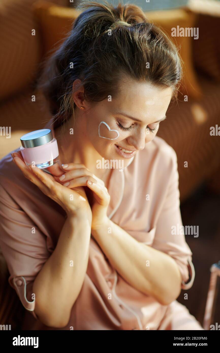 smiling elegant woman in pajamas with white facial cream heart on cheek holding pink cosmetic jar in the modern living room in sunny winter day. Stock Photo