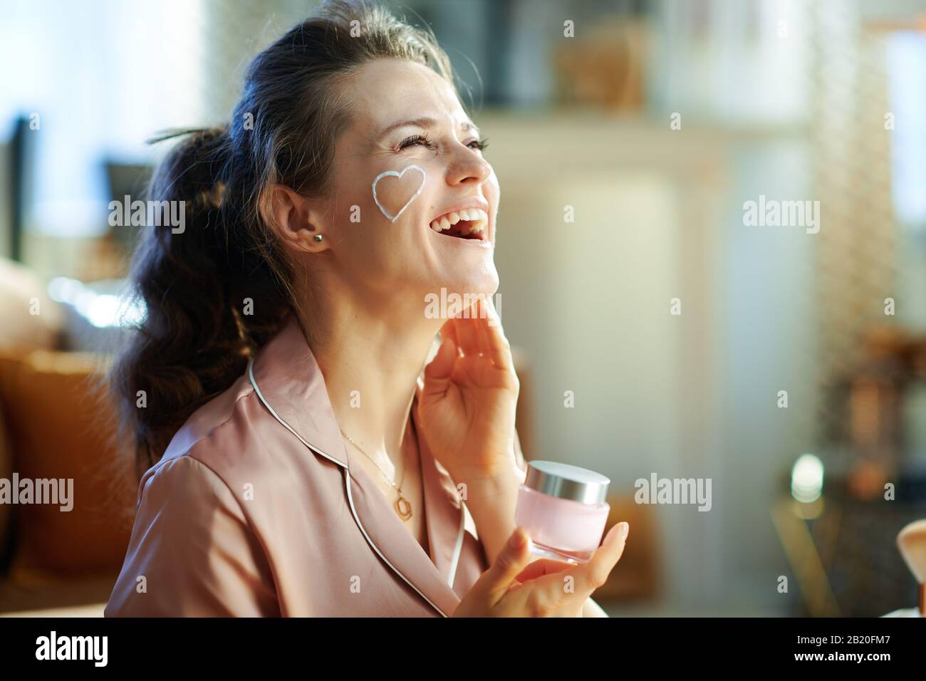 smiling stylish woman in pajamas with white facial cream heart on cheek holding cosmetic jar at modern home in sunny winter day. Stock Photo