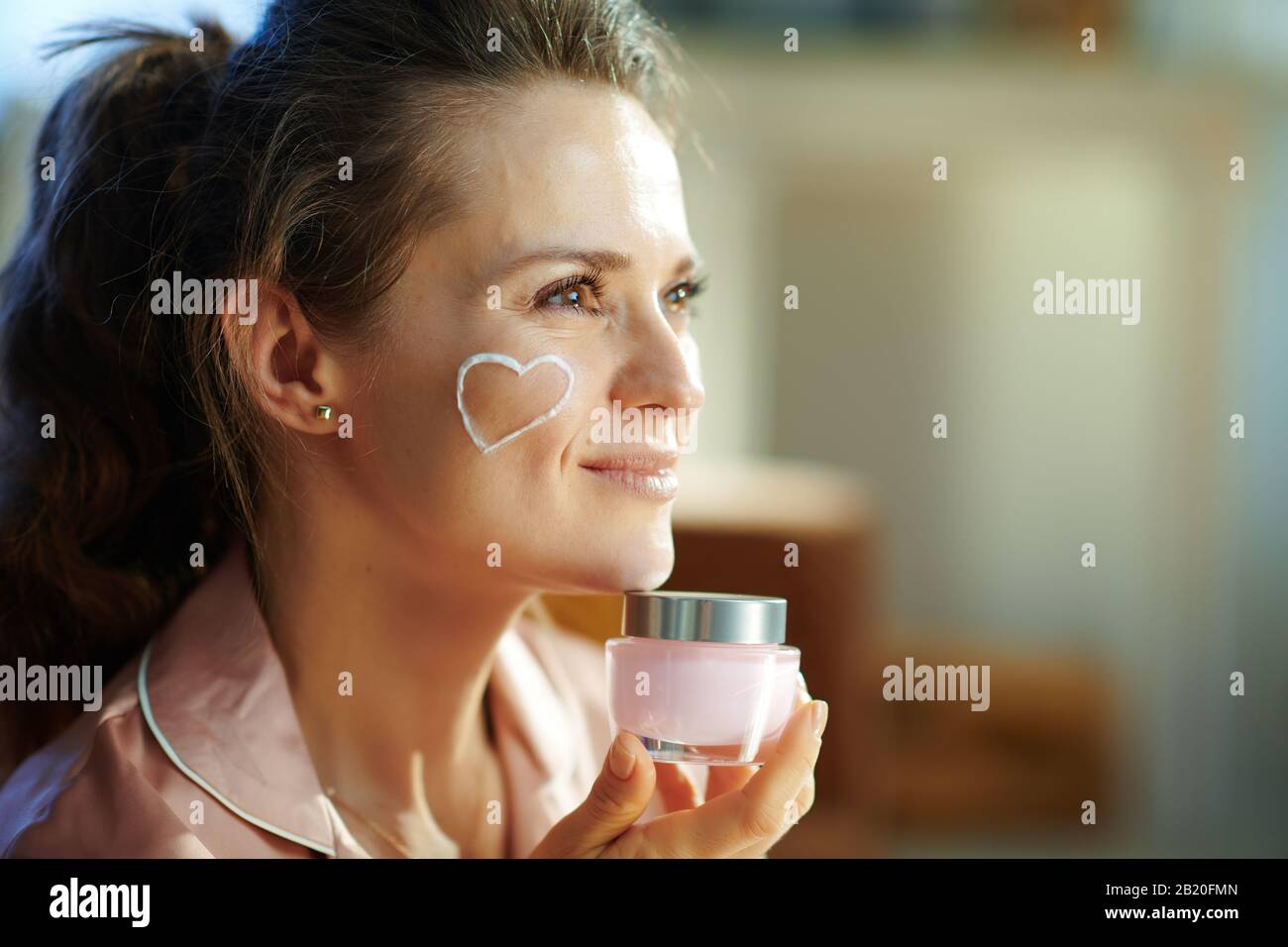 Side view of happy stylish female in pajamas with white facial cream heart on cheek holding pink cosmetic jar at modern home in sunny winter day. Stock Photo