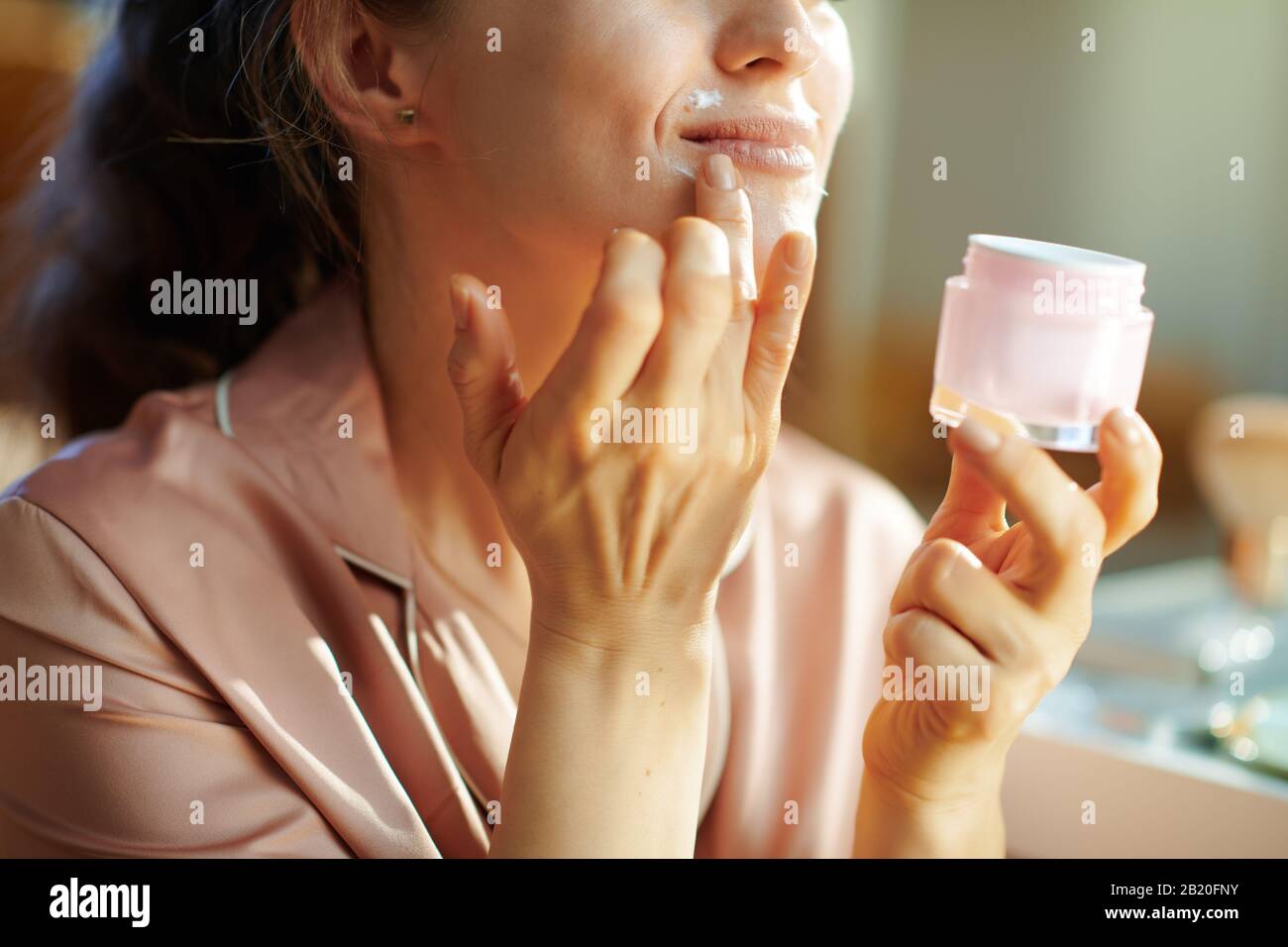 Closeup on happy modern woman in pajamas with pink cosmetic jar applying lip contour cream at modern home in sunny winter day. Stock Photo