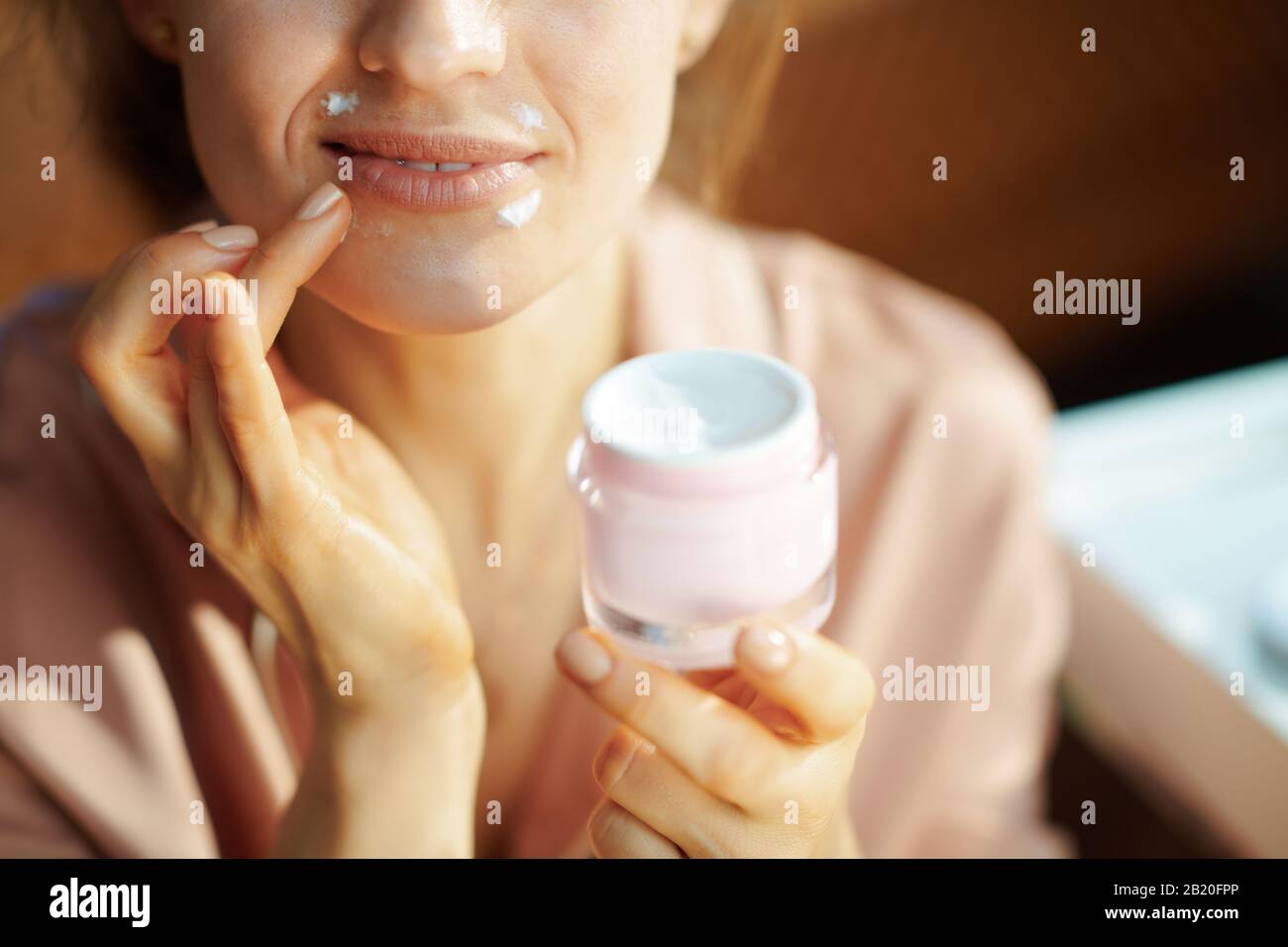 Closeup on smiling elegant woman in pajamas with pink cosmetic jar applying lip contour cream at modern home in sunny winter day. Stock Photo