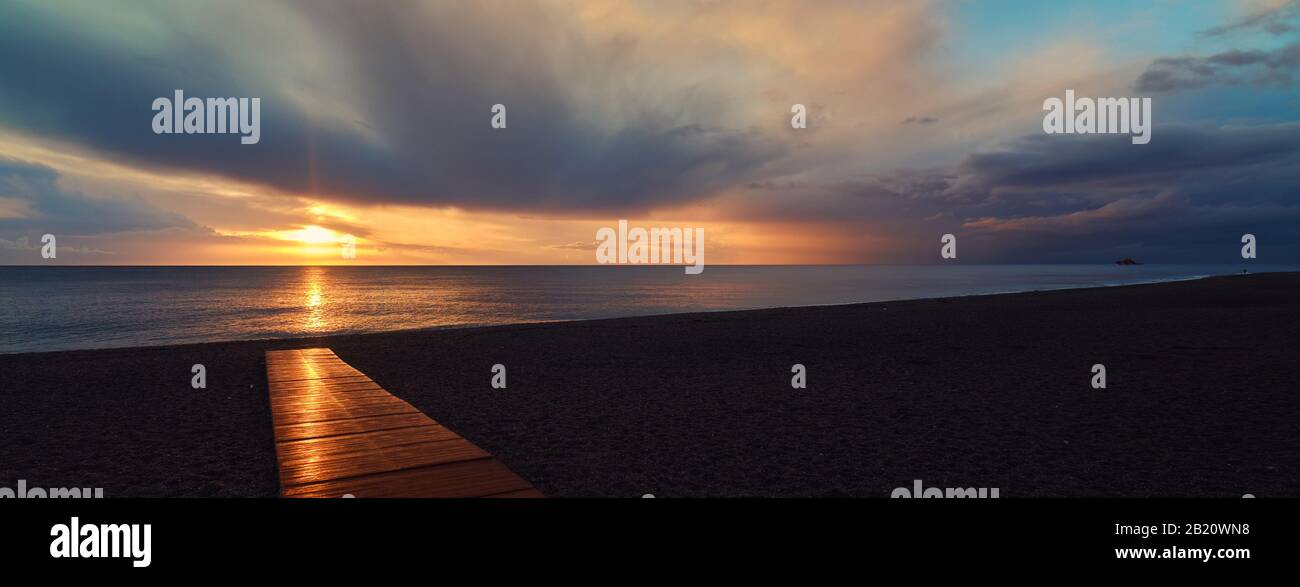 Empty Calahonda beach in Costa del Sol, Andalusia. Bright glowing sun rise beams reflected in wooden boardwalk, fluffy cloudy sky Stock Photo